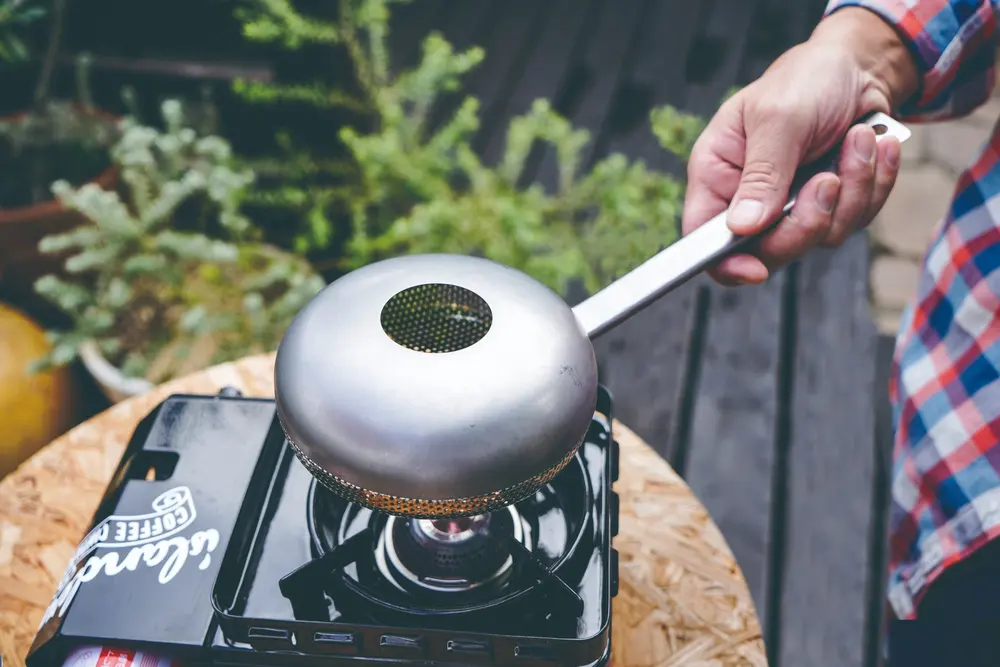 屋外のテーブルで、カセットコンロの炎の上にULTIMATE HAND COFFEE ROASTERをかざして生豆を焙煎している様子
